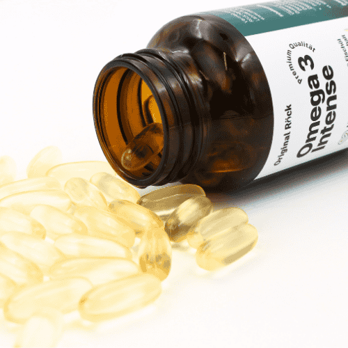 Ein geöffnetes braunes Glas Omega 3 Intense liegt seitlich, während zahlreiche goldene Softgel-Kapseln herausrollen. Die Szene zeigt die glänzende Textur und hochwertige Reinheit der Omega 3 Fischöl Kapseln vor hellem Hintergrund.